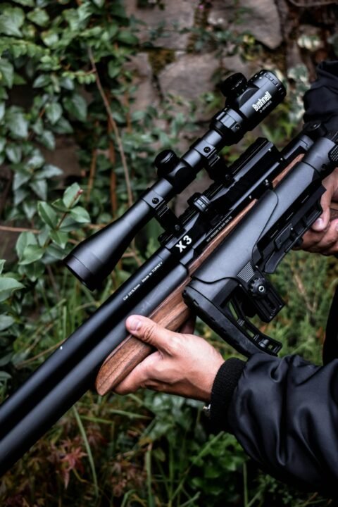 Detailed image of hands holding a scoped rifle against a natural background.